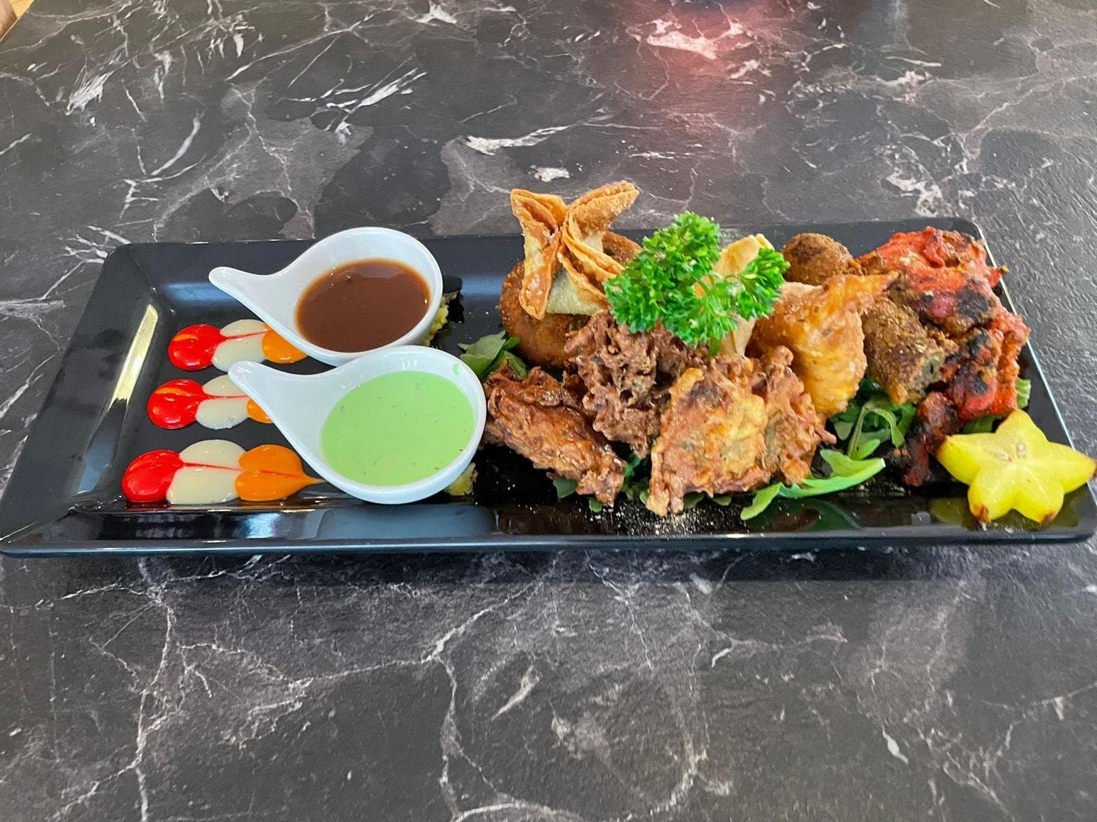 A starter platter of fritters, samosa and tandoori pieces with two dipping sauces, on a black plate.