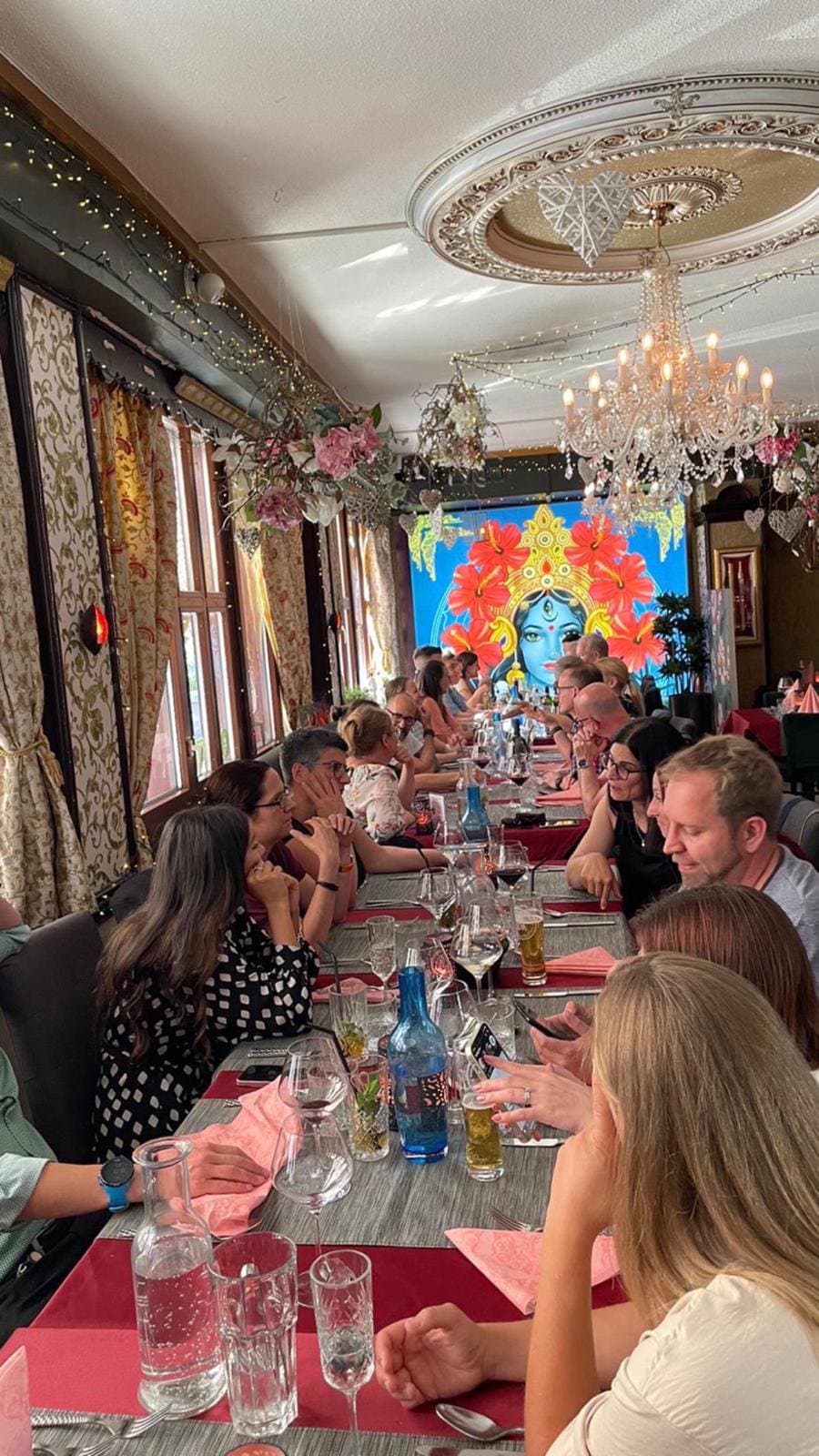 A long table of guests in the dining room, under a crystal chandelier, with a large painted mural at the far end.